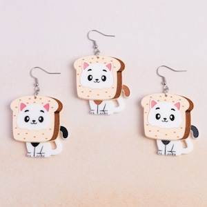 May include: A pair of cat-shaped earrings with a white cat wearing a brown and beige bread costume. The earrings are silver and have a hook closure.
