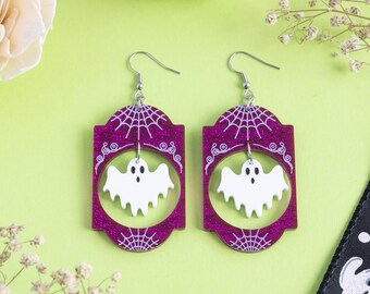 May include: A pair of Halloween-themed earrings. Each earring features a purple, rectangular frame with a spiderweb design and a white ghost cutout. The earrings hang from silver hooks. The background is a light green.