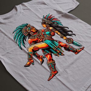 May include: White t-shirt featuring a colorful illustration of two Aztec figures. The male figure is carrying the female figure. The artwork includes teal, red, orange, and gold accents, with detailed headdresses and clothing.