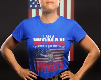 May include: A woman wearing a blue t-shirt with the text "I am a Woman I served in the Military I am a Veteran" and an American flag graphic. She is standing in front of an American flag.