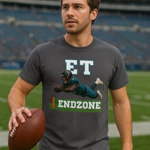 May include: Dark gray t-shirt with the word "ET" in white block letters above an illustration of a football player diving into the endzone. The word "ENDZONE" is printed in green and white below the illustration. The person is holding a brown football.
