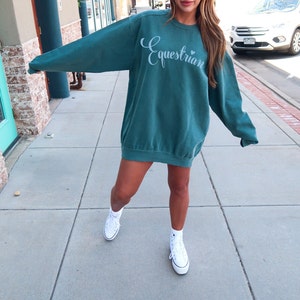 May include: A teal blue sweatshirt with the word "Equestrian" in white script on the front. The sweatshirt is oversized and has a relaxed fit. The model is wearing white sneakers.