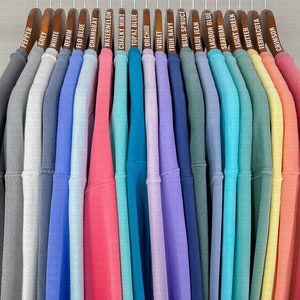 May include: A row of 20 sweatshirts hanging on wooden hangers. The sweatshirts are in various colors, including grey, white, blue, pink, green, yellow, and orange. Each hanger has a label with the color of the sweatshirt written on it.
