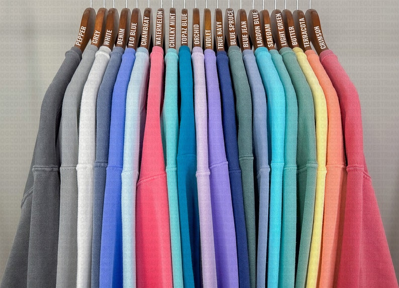 May include: A row of 20 sweatshirts hanging on wooden hangers. The sweatshirts are in various colors, including grey, white, blue, pink, green, yellow, and orange. Each hanger has a label with the color of the sweatshirt written on it.