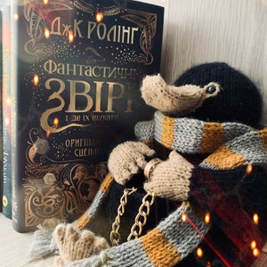 May include: A knitted black and brown Niffler plush toy wearing a gray and orange striped scarf and a gold chain. The toy is sitting next to a book titled "Fantastic Beasts and Where to Find Them" by J.K. Rowling.