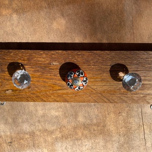 May include: A wooden wall-mounted coat rack with three hooks. Two hooks are clear glass knobs and one hook is a ceramic knob with a black and orange floral design.