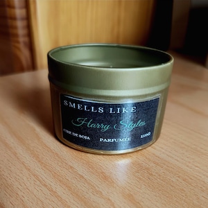 Peut inclure: Une bougie en pot doré avec une étiquette noire indiquant "Smells Like Harry Styles". La bougie est faite de cire de soja et pèse 110g. Le pot est posé sur une surface en bois.