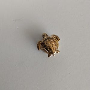 Peut inclure: Une petite broche en métal doré en forme de tortue de mer. La tortue présente un motif de carapace détaillé et en relief, avec une finition lisse et réfléchissante sur la tête et les nageoires, reproduisant l'anatomie de l'animal marin.