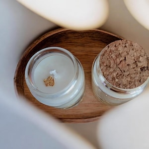 Peut inclure: Deux petits bocaux en verre contenant de la cire blanche, l'un ouvert avec une mèche blanche et un éclat doré, l'autre fermé par un bouchon en liège rond. Les deux bocaux sont posés sur un plateau en bois ovale au grain naturel visible.