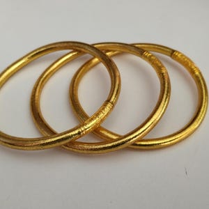 Pode incluir: Três pulseiras douradas entrelaçadas, criando um efeito em camadas. As pulseiras têm um design liso e arredondado com textura sutil e acabamento brilhante. As joias são exibidas em um fundo branco liso.