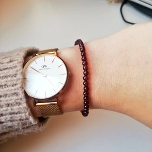 Peut inclure: Une montre dorée avec un cadran blanc et un bracelet en maille. La montre porte l'inscription "DW Daniel Wellington" sur le cadran. La montre est portée au poignet avec un bracelet de perles rouges.