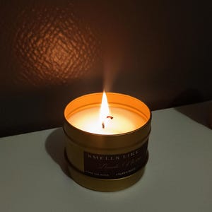 Puede incluir: Un tarro de vela dorado con una vela de cera de soja blanca en su interior. La vela est&aacute; encendida y la llama es visible. La etiqueta del tarro dice "Smells Like" y "Lumberjack" en una fuente cursiva. Tambi&eacute;n se puede ver el texto "Cire de Soja" y "Parfum&eacute;e" en la etiqueta.