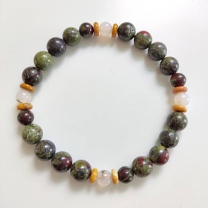 May include: A beaded bracelet featuring a mix of round, natural stone beads in shades of green, burgundy, and tan. The bracelet also includes smaller, lighter-colored beads, creating a unique and earthy accessory.