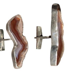 May include: A pair of cufflinks featuring a natural agate stone with brown and white bands, set in a silver-toned metal. The cufflinks have a unique, organic shape, and a classic T-bar closure.