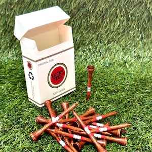 May include: A collection of wooden golf tees with red and white stripes, scattered on artificial green grass. A white box with the text "LUCKY TEES" and "ORIGINAL RED" is open in the background. The tees are designed for 300+ yards.