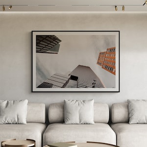 May include: A modern living room with a large sectional sofa, two round coffee tables, and a single armchair. The sofa is light gray with white pillows. The coffee tables are brown with a wood top. The armchair is light gray with a black metal frame. The room has a white ceiling with track lighting and a large framed photograph of city buildings on the wall.