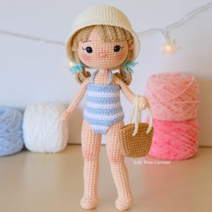 Puede incluir: Muñeca de ganchillo con sombrero blanco y traje de baño a rayas azules y blancas, sosteniendo una pequeña bolsa tejida. La muñeca tiene cabello rubio y está de pie frente a ovillos de lana. La imagen incluye el texto "PDF pattern instant download" y una bandera estadounidense.