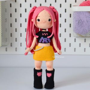 May include: A handmade crochet doll with pink hair in pigtails, wearing a black top with a purple and blue design, a yellow skirt, and black boots. The doll is standing in front of a white pegboard. Text on the left says "PDF pattern instant download" with an American flag.