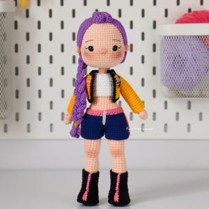 Puede incluir: Muñeca de ganchillo con una trenza morada, que lleva una chaqueta amarilla y negra, pantalones cortos azules y botas negras. La muñeca tiene tez clara y está de pie frente a una placa perforada blanca. El texto de la imagen dice "PDF pattern instant download".