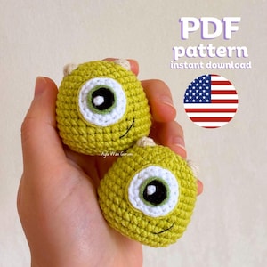 May include: Two crocheted green and yellow amigurumi toys with one large eye and a small mouth. The toys are in the shape of a monster with a single eye. The toys are being held in a person's hand. PDF pattern instant download.