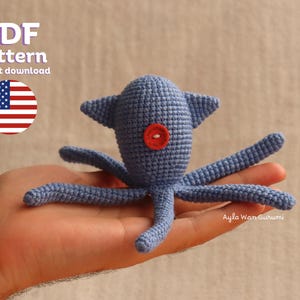 May include: A blue crocheted octopus amigurumi toy with a red button for an eye. The toy is being held in a person's hand. The text "Ayla Wamigurumi" is visible in the bottom right corner of the image.