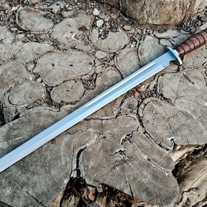 May include: A silver-bladed sword with a brown leather-wrapped handle. The blade is straight and tapers to a point. The handle is cylindrical and appears to be made of stacked leather. The sword rests on a weathered, textured wooden surface.