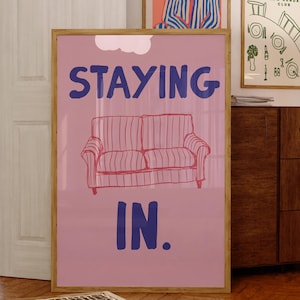 Puede incluir: Un póster rosa enmarcado con las palabras "STAYING IN" en azul, encima de una ilustración roja y rosa de un sofá. El póster está en una habitación con otras obras de arte enmarcadas y muebles de madera.