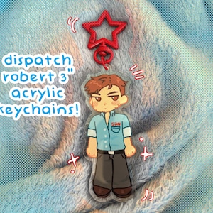 Puede incluir: Llavero acrílico con una ilustración de dibujos animados de un personaje con camisa azul y pantalón gris. El llavero tiene un cierre rojo en forma de estrella y dice "dispatch robert 3" en texto azul. El fondo es una tela azul claro texturizada.