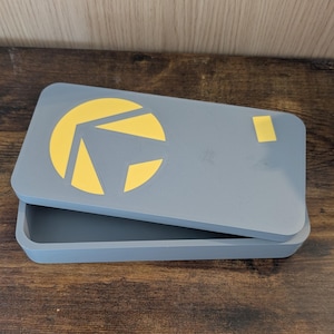 Puede incluir: Una caja rectangular gris con tapa, con un diseño geométrico amarillo. La tapa está ligeramente entreabierta, revelando el interior de la caja. La caja está sobre una superficie de madera.