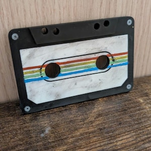 Puede incluir: Una cinta de cassette en blanco y negro con un diseño retro. La cinta presenta una etiqueta blanca con rayas coloridas rojas, naranjas, verdes y azules. La cinta de cassette es un artículo nostálgico.