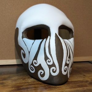 Könnte beinhalten: Eine weiße Keramikmaske mit braunen Details. Die Maske hat zwei Augenlöcher und ein Design aus wirbelnden Linien auf der unteren Hälfte.