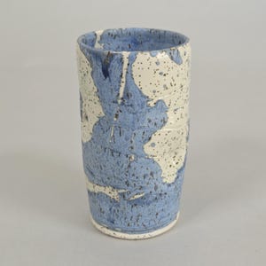 May include: A cylindrical ceramic tumbler featuring a speckled cream-colored base with irregular, flowing patches of blue glaze. The surface has a textured appearance with visible horizontal ridges along the body and a smooth, glossy finish.