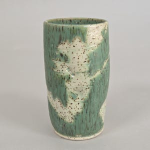 May include: A cylindrical ceramic tumbler featuring a reactive glaze with overlapping layers of sage green and off-white. The surface displays a speckled texture with dark brown flecks distributed across the light-colored areas.