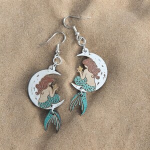 May include: A pair of silver earrings with a crescent moon design. Each moon features a mermaid holding a star, with a teal tail dangling below.