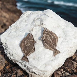 May include: A pair of wooden earrings shaped like halibut fish. The earrings are brown and have a natural wood grain finish. They are hanging from silver hooks.
