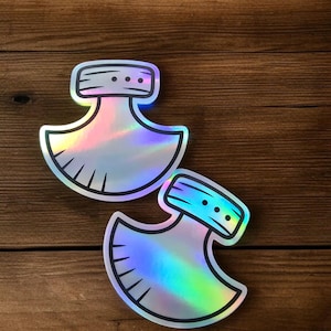 Op de afbeelding: Twee holografische stickers met een uniek, bijlachtig ontwerp. Elke sticker heeft een zilveren omtrek, een rechthoekige bovenkant met drie stippen en een gebogen mesvorm. De stickers hebben een regenboogkleurige iriserende afwerking.