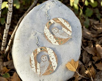 May include: A pair of wooden earrings featuring a white owl perched in a birch tree design. The earrings are round and have a silver hook.