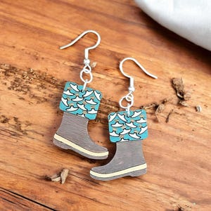 May include: A pair of wooden earrings shaped like rain boots. The boots are brown with a teal and white patterned top. The earrings have silver hooks.
