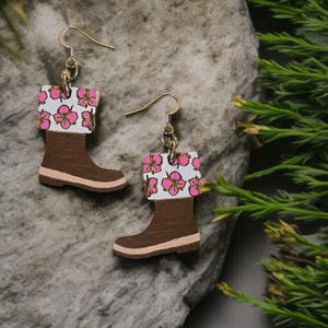 May include: A pair of wooden earrings shaped like brown rain boots with a white cuff adorned with pink flowers. The earrings have gold-colored ear wires and are set against a gray stone background with green foliage.