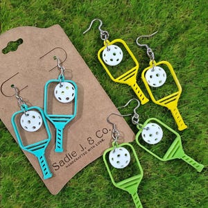 May include: A pair of pickleball paddle earrings. The earrings are made of acrylic and feature a white pickleball with black holes. The paddles are available in yellow, green, and turquoise.