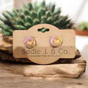 May include: A pair of wooden earrings with a pink, orange, and yellow sunset design. The earrings are on a brown card with the text "Sadie J. & Co. Handcrafted with Love".