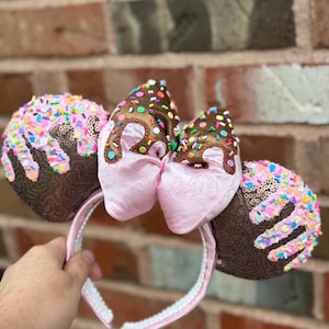 May include: A pink headband with two chocolate-covered ears and a pink bow. The ears are brown with pink sprinkles and a chocolate drizzle. The bow is pink with a chocolate drizzle and sprinkles. The headband is white with pink trim.