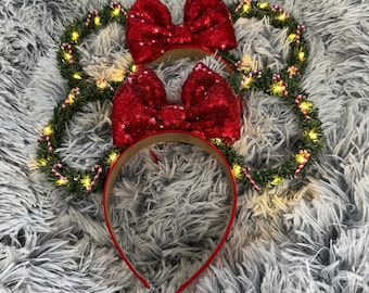 May include: Red sequined bow Minnie Mouse ears with a green garland and white lights. The garland is decorated with candy canes.