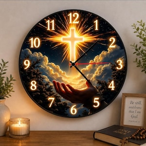 May include: A round wall clock with a black frame. The clock face features a glowing cross, clouds, and a hand reaching upwards. The numbers are in a glowing font. The clock is on a wooden surface with a candle and a framed quote.