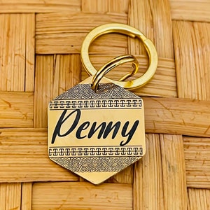 May include: A gold-coloured, hexagonal pet tag with the name "Penny" in a stylish script. The tag is attached to a gold keyring. The background is a woven, light brown surface.