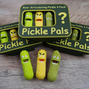 Könnte beinhalten: Drei grüne und gelbe, bewegliche Gurkenspielzeuge mit ausdrucksstarken Gesichtern, zusammen mit drei grünen und gelben Schachteln mit der Aufschrift "Sour Articulating Pickle 4 Pack" und "Pickle Pals". Die Schachteln haben ein Fragezeichen.