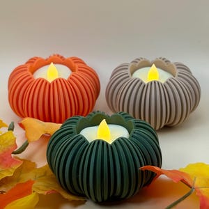Può includere: Tre portacandele a forma di zucca in arancione, beige e verde scuro, ognuno caratterizzato da una consistenza a coste e un lumino LED bianco centrale con una fiamma gialla brillante, accompagnati da foglie autunnali artificiali sparse.