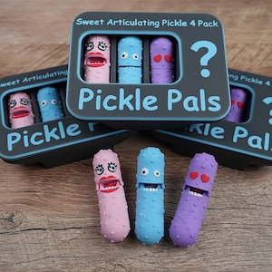 May include: A set of three colorful, articulated pickle-shaped toys with faces, alongside boxes labeled "Pickle Pals." The pickles are pink, blue, and purple, each with unique facial features. The boxes are dark gray with the text "Sweet Articulating Pickle 4 Pack."