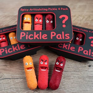 May include: A set of three articulated pickle toys in orange, red, and dark red, with angry facial expressions. The toys are displayed with three black boxes with the text "Pickle Pals" and "Spicy Articulating Pickle 4 Pack" in red.
