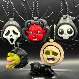 May include: Five horror-themed keychains featuring a white ghost mask, a red devil, a skull in a black hood, a yellow hand with pink fingernails, and a beige mummy head. Each charm includes a metal ball chain for attachment and detailed textures.